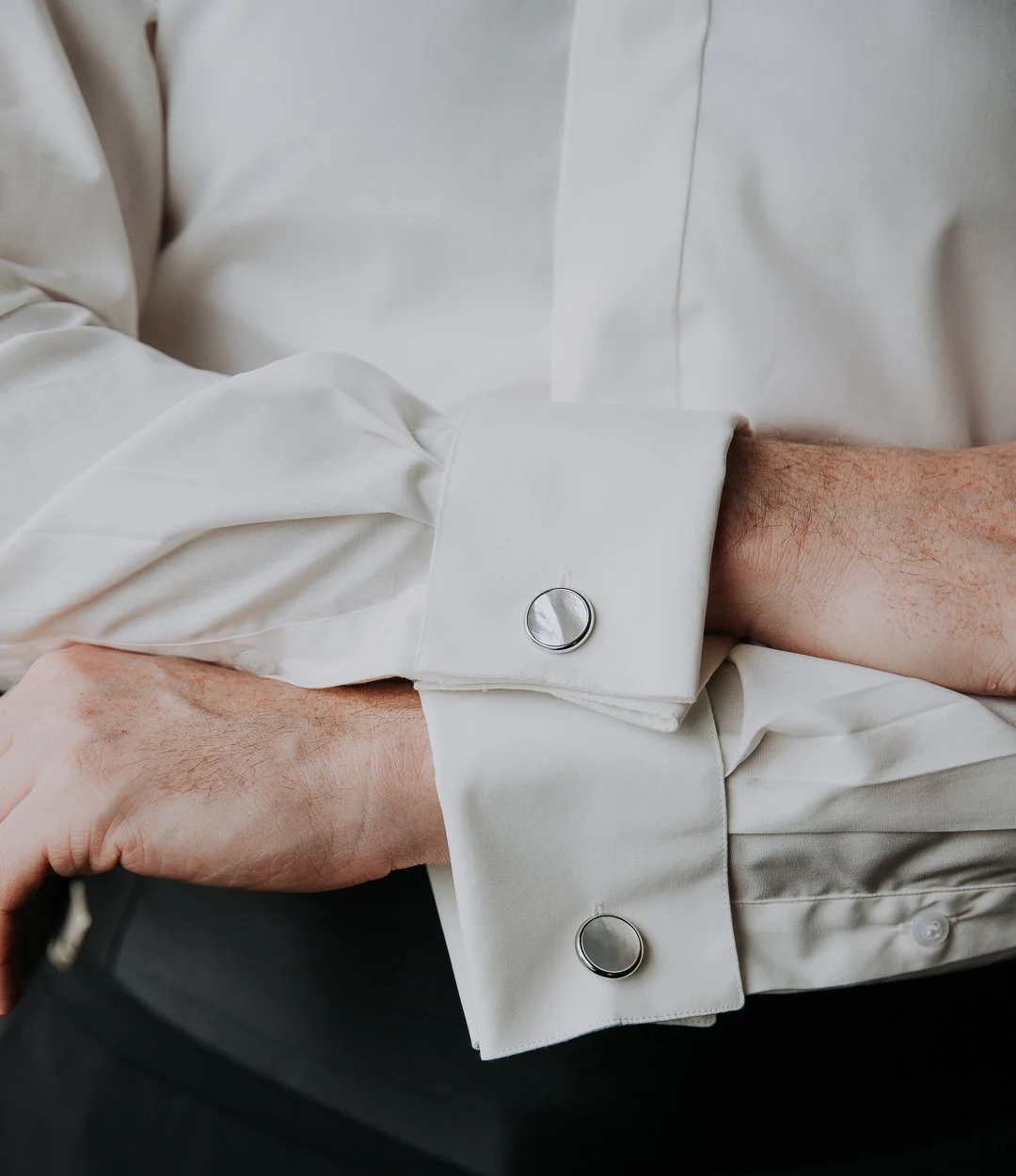 Close-up of a person wearing a white dress shirt with silver cufflinks on the cuffs, with hands resting on a surface.