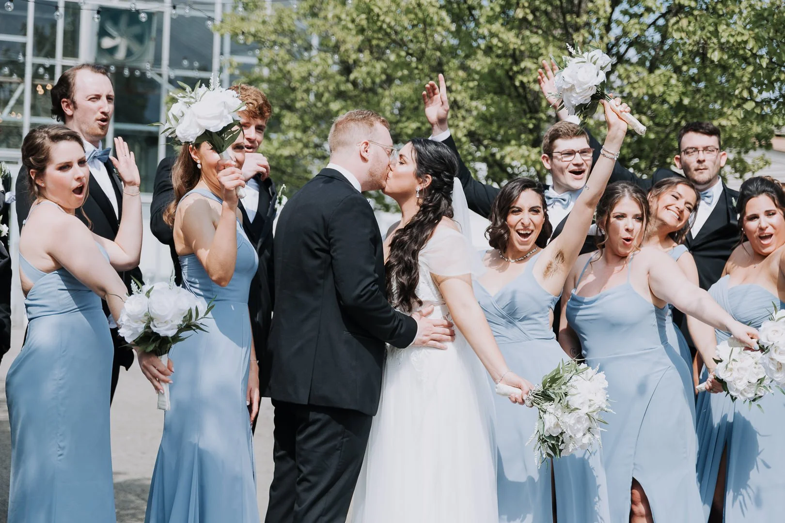 A wedding party outdoors with the bride and groom kissing, surrounded by bridesmaids in light blue dresses holding bouquets, and groomsmen in black tuxedos with light blue bow ties, celebrating and smiling.