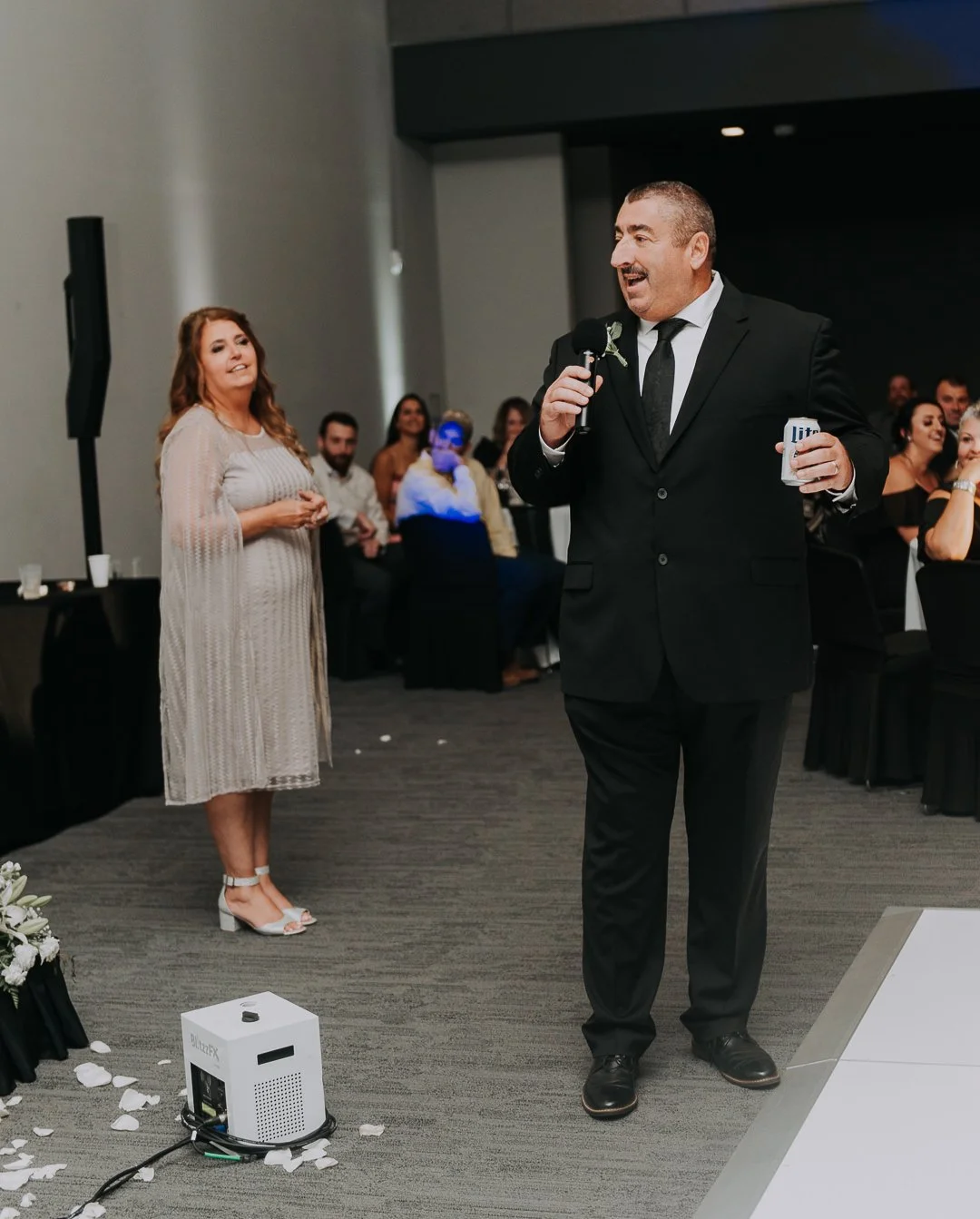A man in a black suit giving a speech at a wedding reception, holding a microphone and a beer can, with a woman in a light dress listening and smiling in the background.