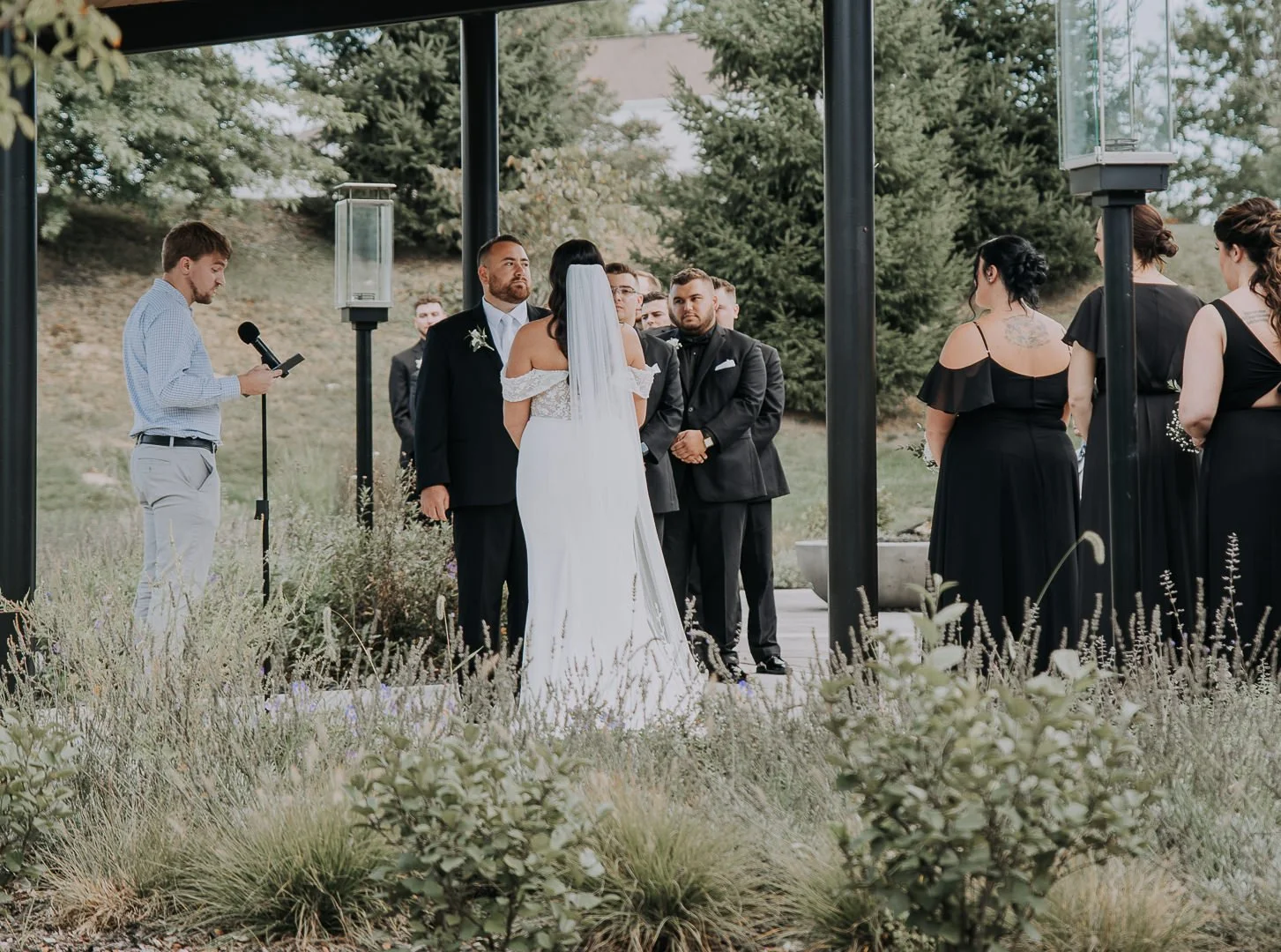 A outdoor wedding ceremony with a bride and groom standing before officiant and guests, surrounded by lush greenery.