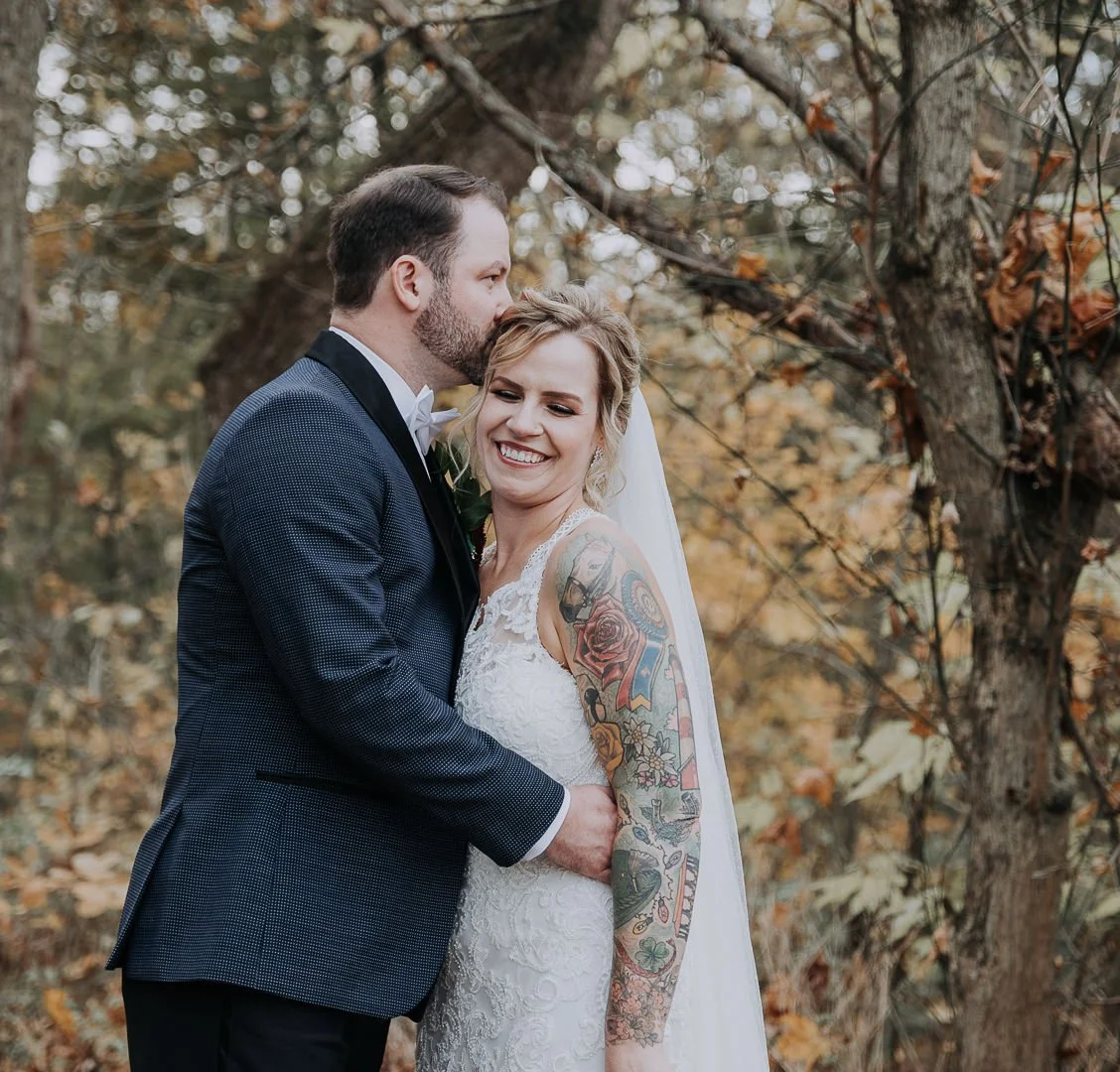 A bride and groom smiling and embracing outdoors in a wooded area with autumn leaves, the groom kissing the bride's forehead and the bride showing her tattooed arm.