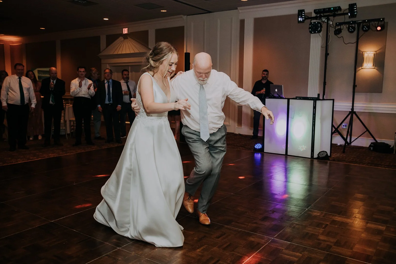 A bride and an older man dance together in a wedding reception hall, surrounded by seated guests and a DJ setup with colorful lights.