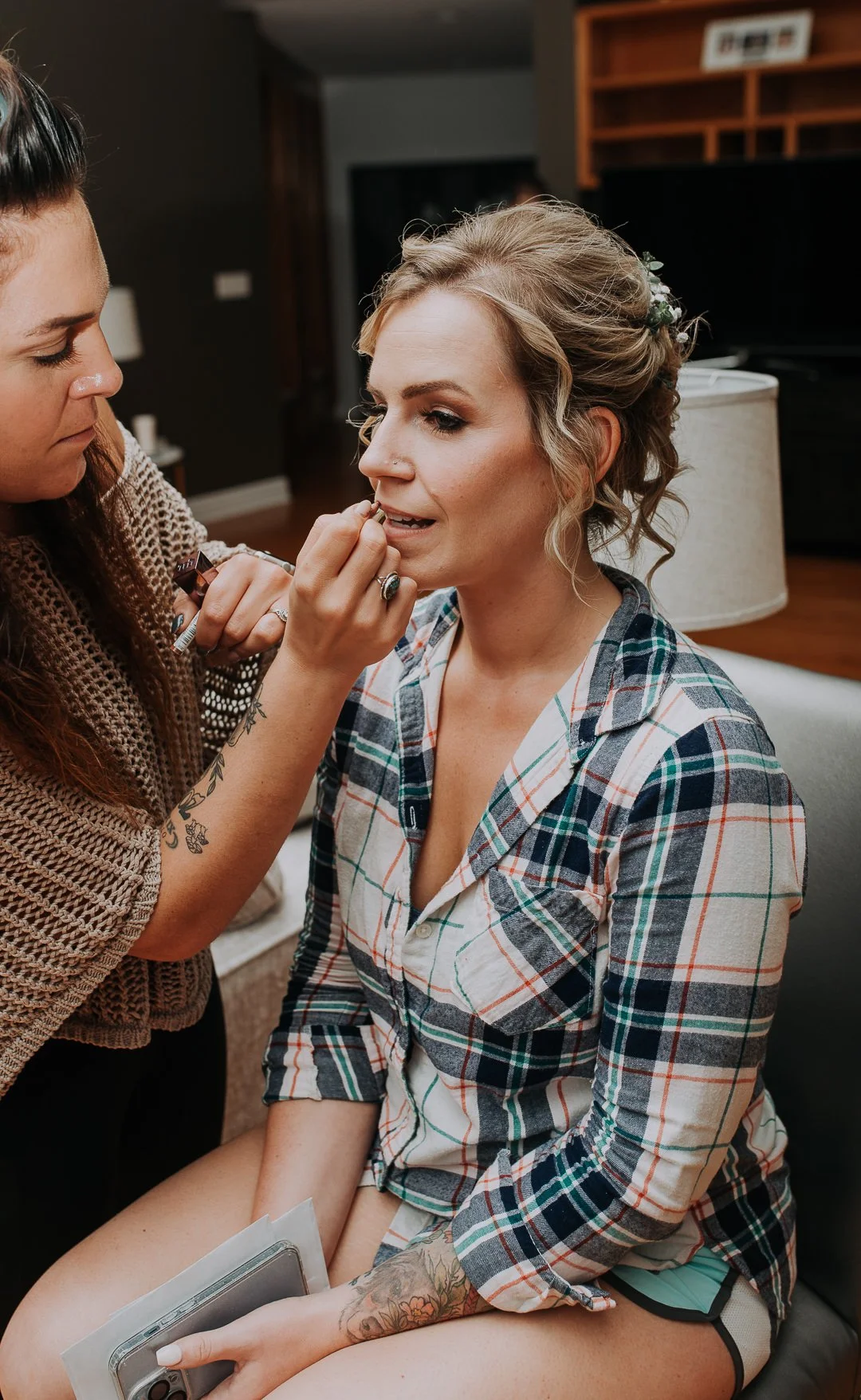 A woman with blonde hair sitting on a chair, getting her lips done by a makeup artist or aesthetician. The woman is wearing a plaid shirt and has tattoos on her arm, while the makeup artist is focused on her task.