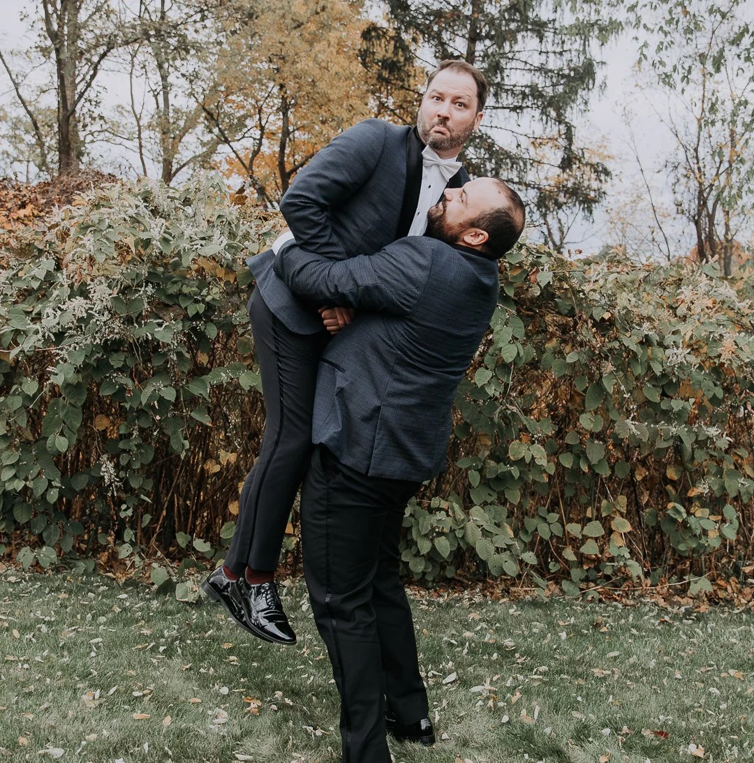 Two men in formal suits outdoors, one lifting the other in an embrace, surrounded by trees and bushes.