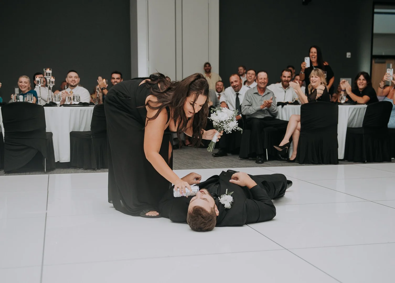 A woman is kneeling and pouring a drink on a man lying on the floor at a wedding reception, with guests clapping and taking photos in the background.