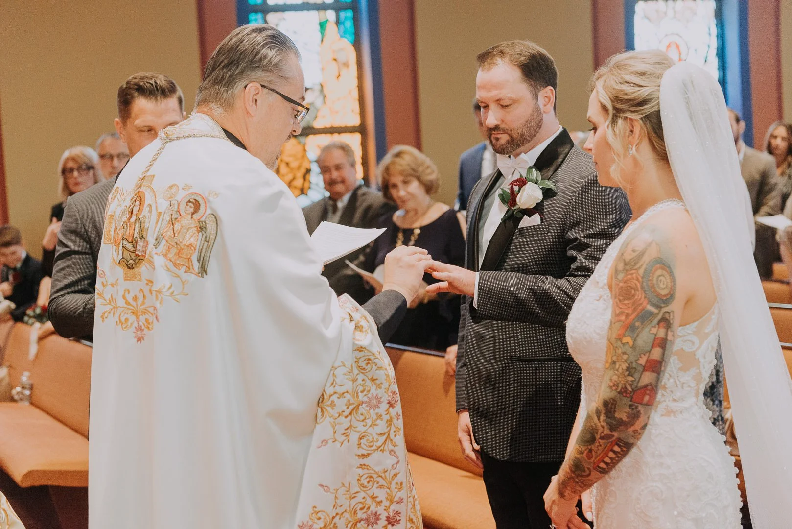 A wedding ceremony inside a church with a priest officiating. The groom, wearing a black tuxedo with a rose boutonniere, and the bride, wearing a white lace wedding dress with colorful tattoos on her arm, are exchanging rings. The background shows we