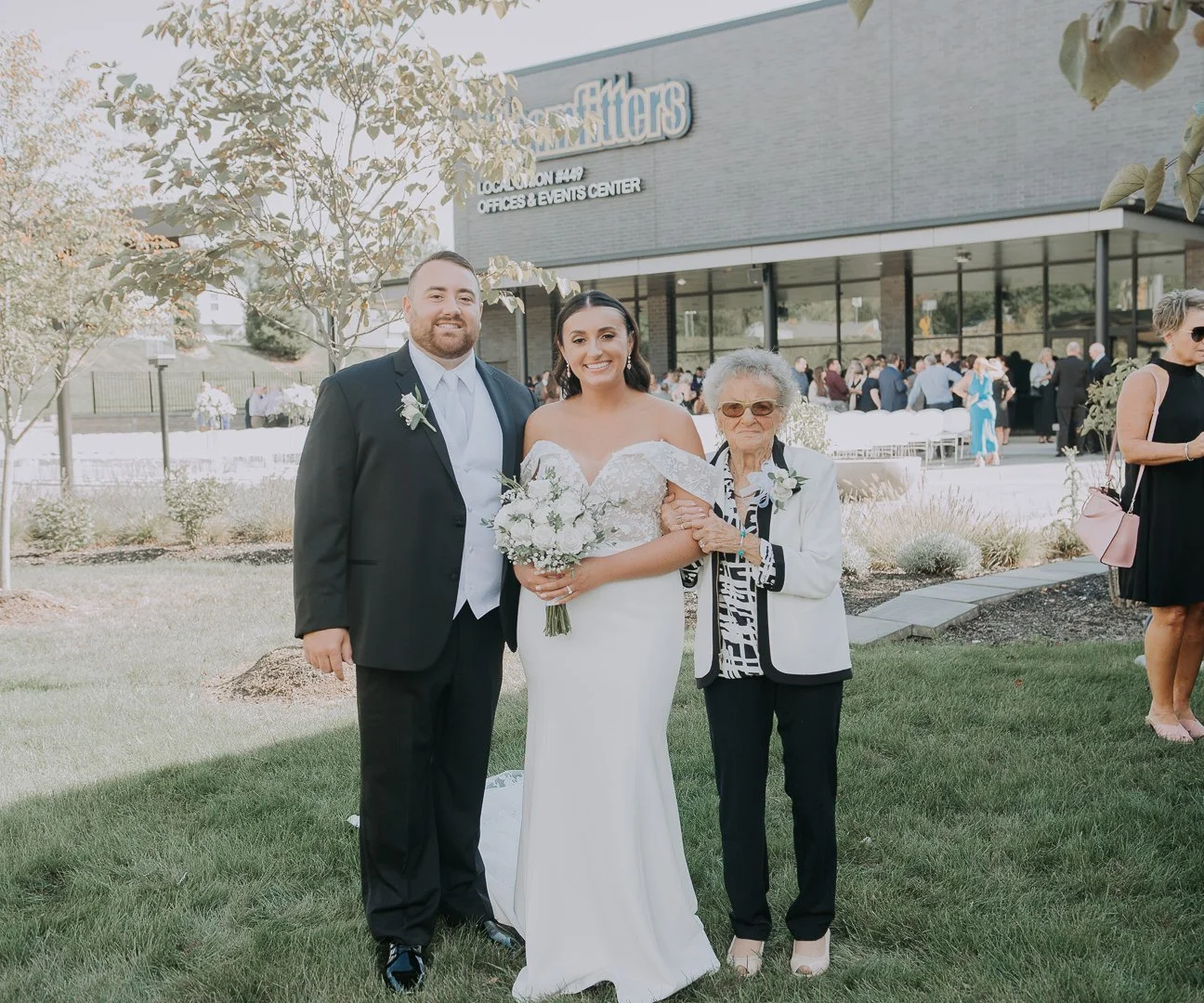 A bride in a white wedding dress holding a bouquet of white flowers, standing between a groom in a black suit and an elderly woman in a white blazer and black pants, outside a modern event center with many guests in the background.