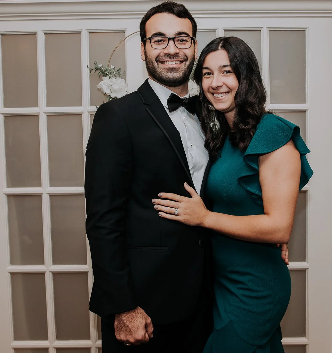 A man and woman are dressed formally, smiling for a photo at an event. The man is wearing a black tuxedo with a bow tie and glasses, and the woman is wearing a teal dress with ruffled sleeves. They are standing in front of a white paneled wall with a