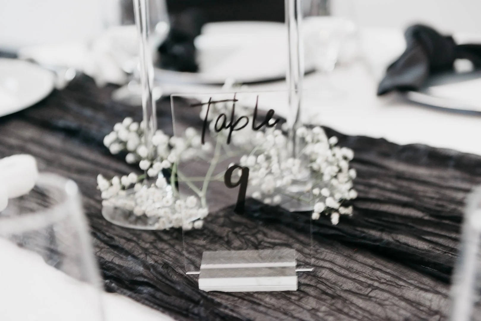 Table centerpiece with a glass sign reading 'Table 9', baby's breath flowers, and a black table runner on a white table setting.