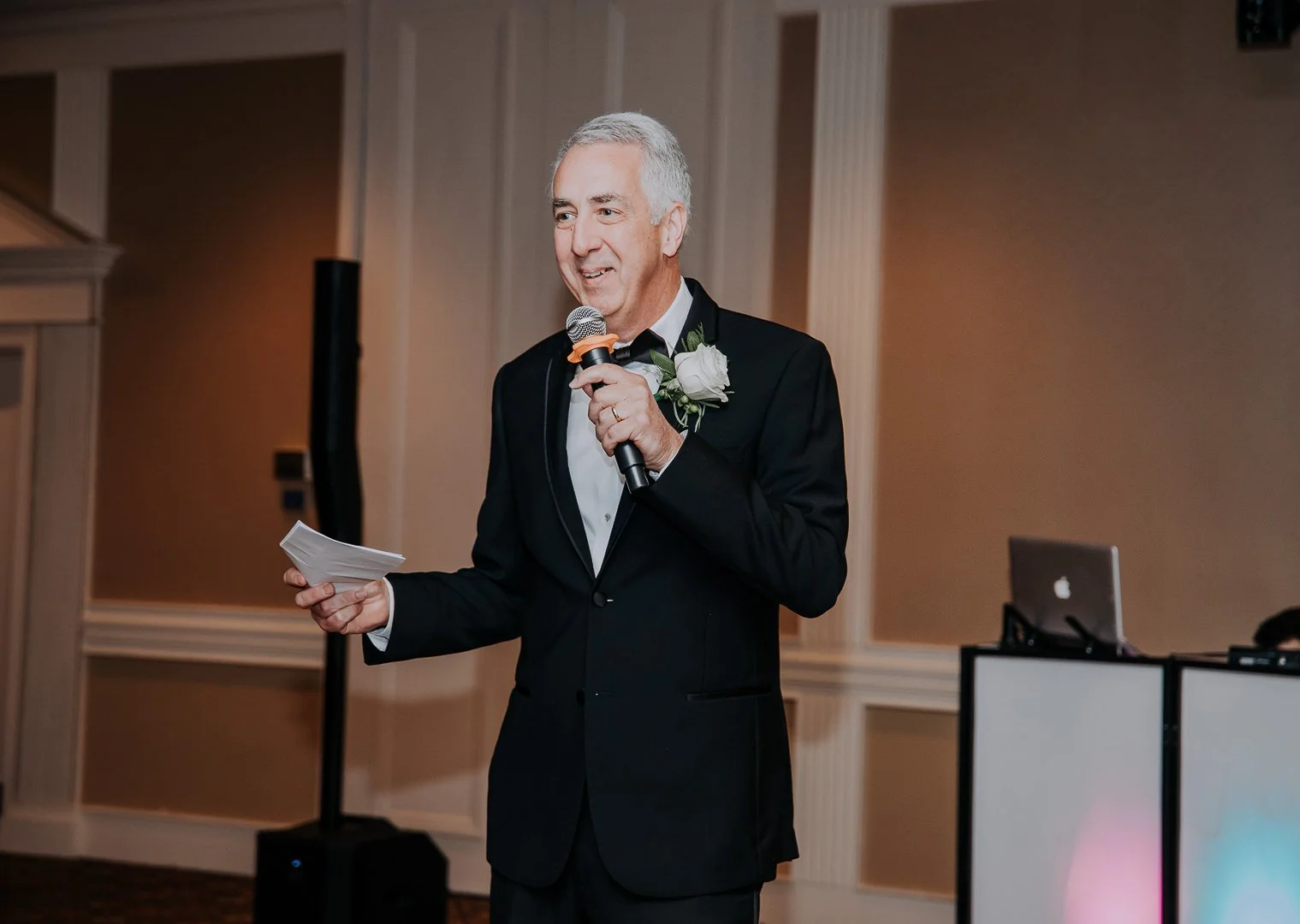 Older man in a tuxedo giving a speech at a formal event, holding a microphone and notes, with a white flower boutonniere on his lapel.