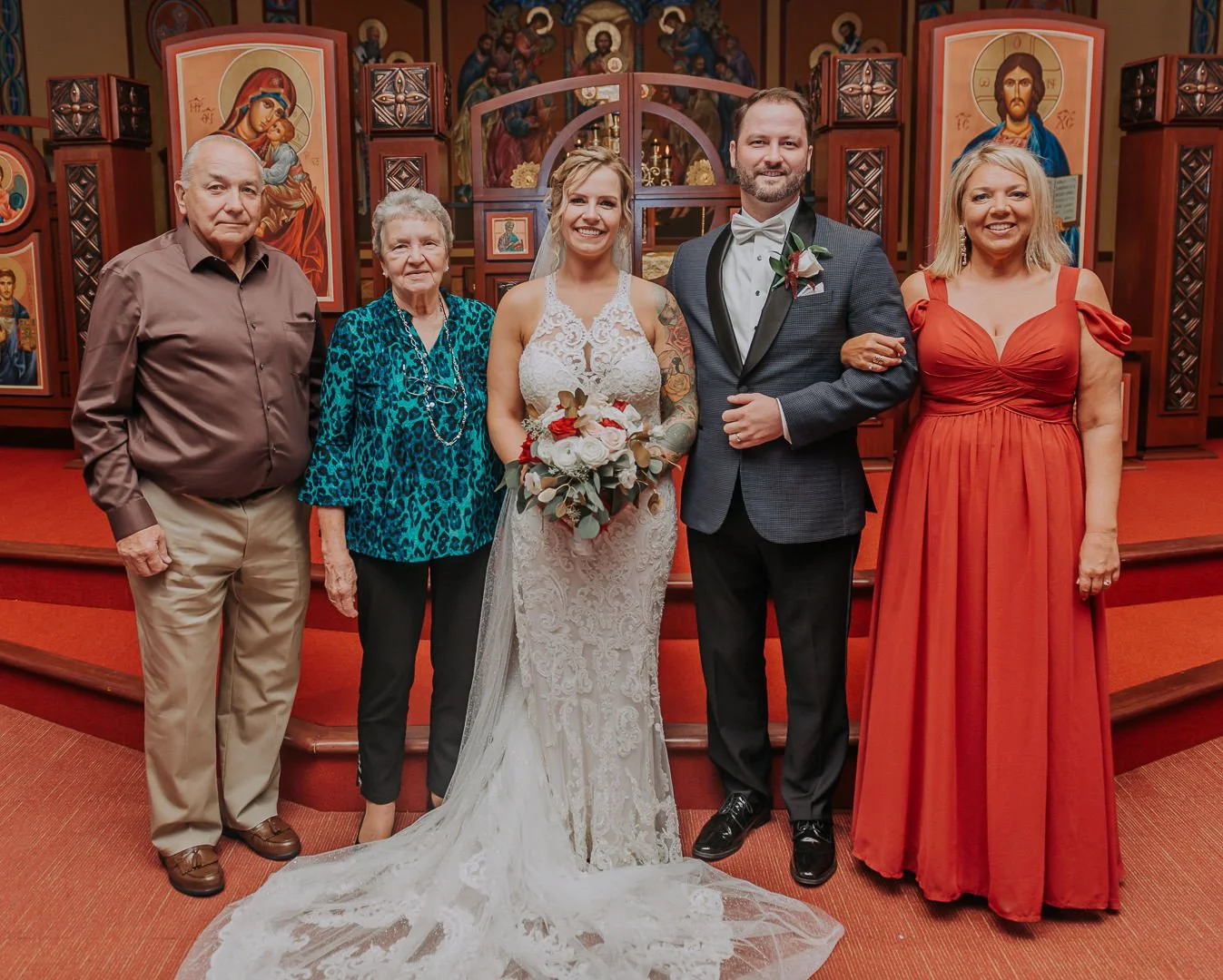 A bride and groom standing arm-in-arm with four family members inside a church, with religious icons in the background. The bride is wearing a white lace wedding dress and holding a bouquet, and the groom is in a gray tuxedo with a bow tie. The famil
