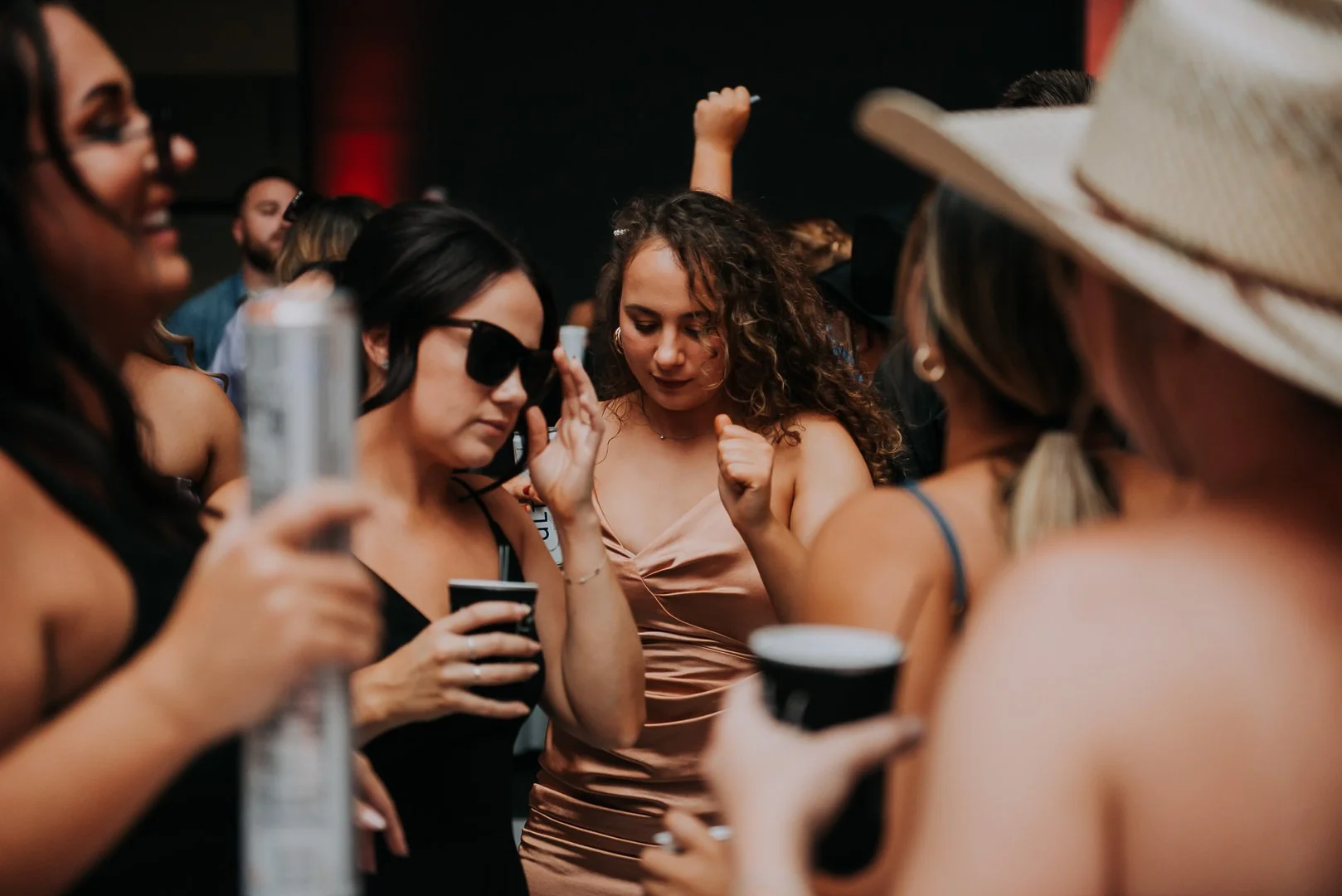 Group of women dancing at a party, holding drinks, in a dark room with red lighting.