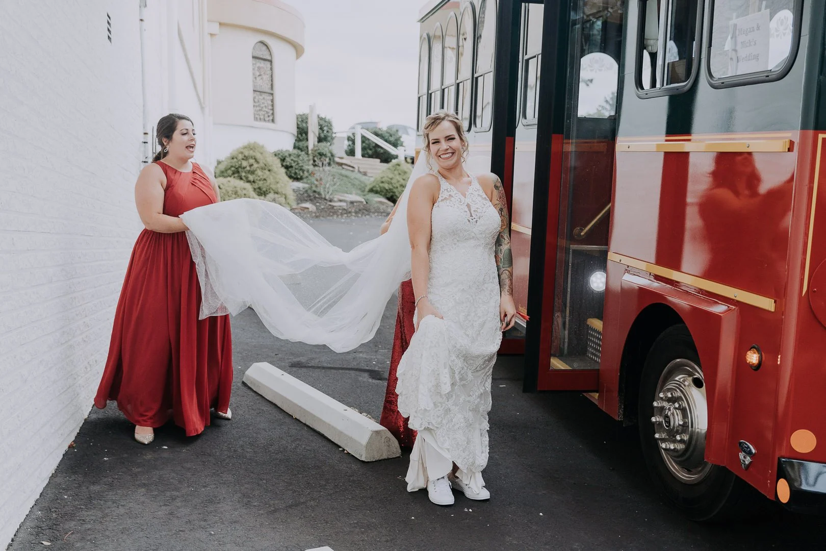 A bride in a white lace wedding dress and sneakers smiling as she steps off a red trolley bus, with a bridesmaid in a red dress holding her train and laughing outside a white building.