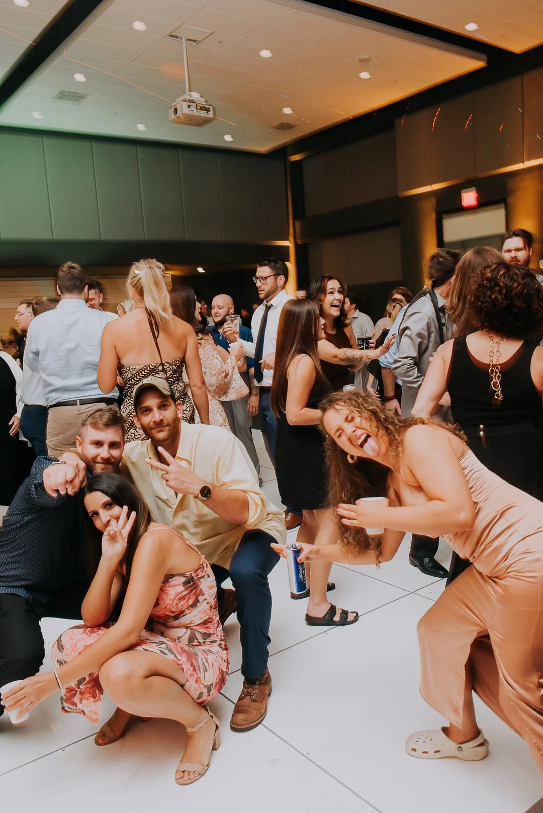 Group of people at a party dancing and socializing in an indoor venue with a dark ceiling and a projector mounted on the ceiling.