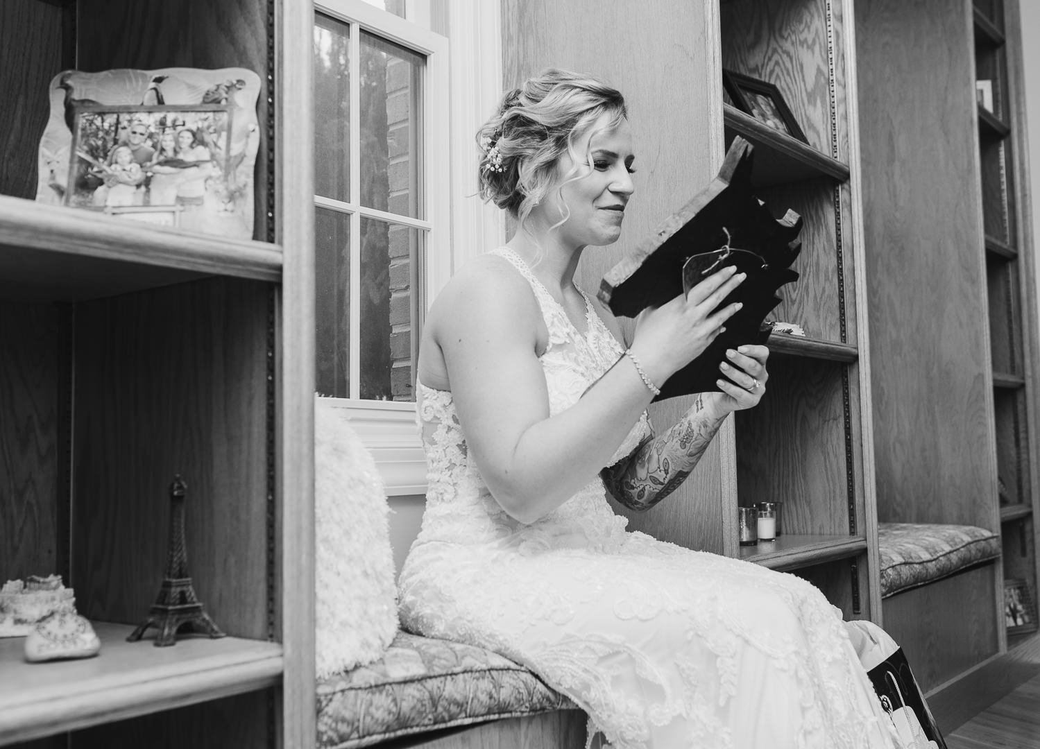 A woman in a wedding dress sitting on a bench reading a card inside a room with wooden shelves and framed photographs.