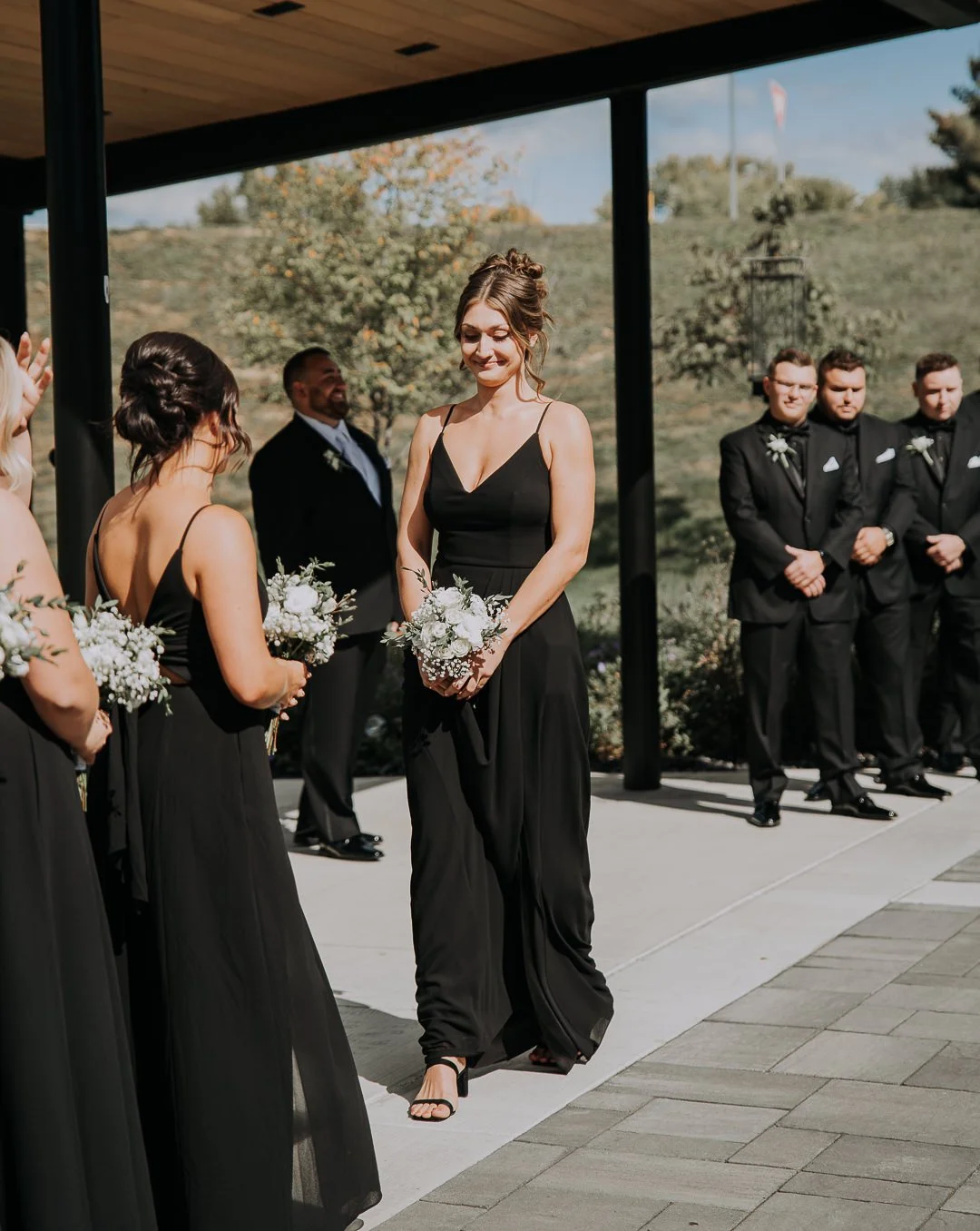 A woman in a black dress holding a bouquet of flowers, standing with other women in black dresses during a wedding ceremony outdoors with groomsmen in black suits in the background.