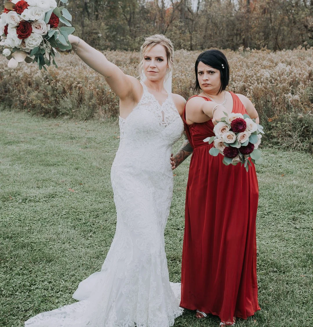 Two women standing outdoors in a grassy field, one in a white wedding dress holding a bouquet, the other in a red dress holding a bouquet, both making assertive gestures.