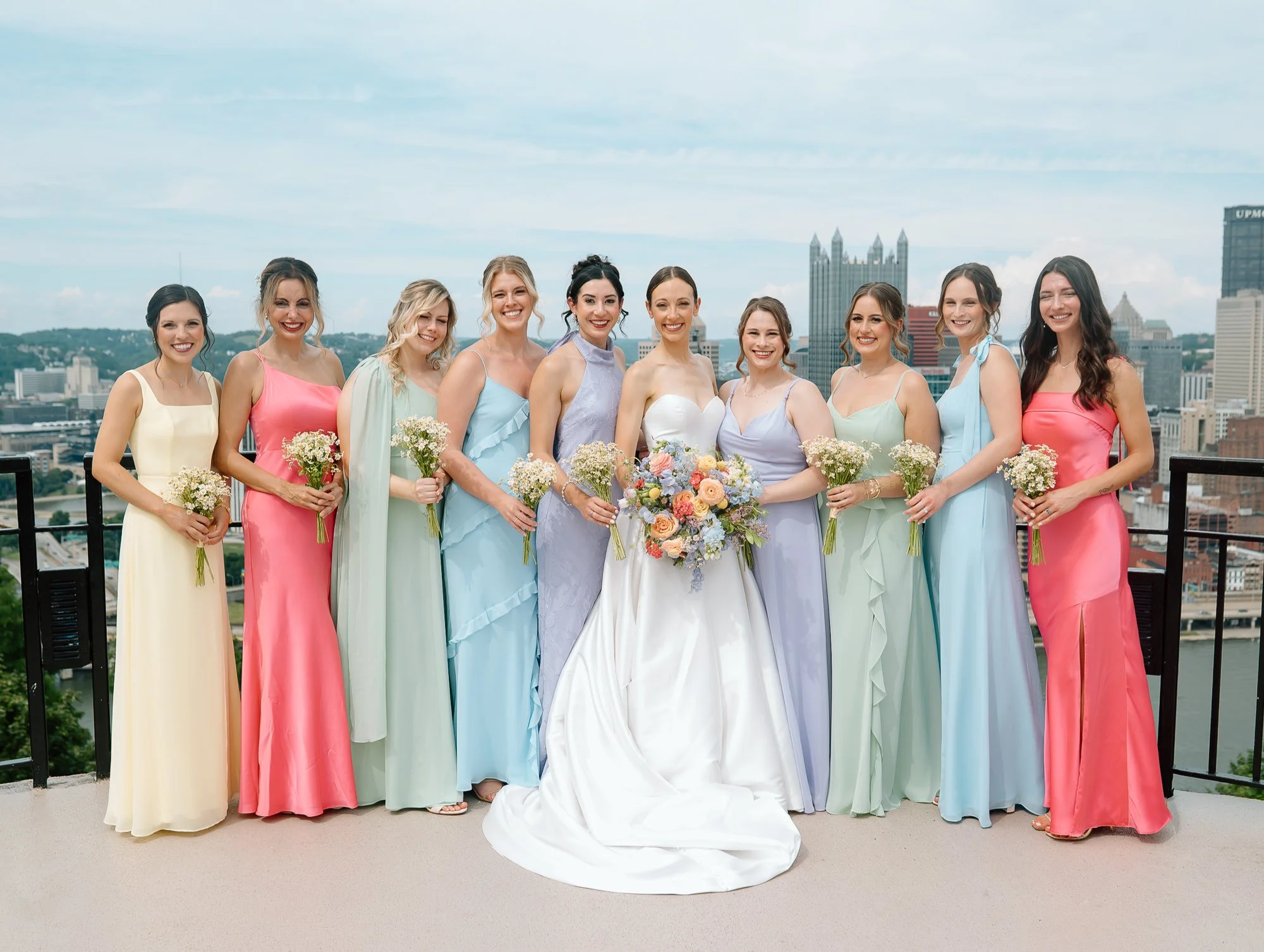 A group of eleven women in pastel dresses standing on a rooftop with a city skyline in the background, holding flowers, smiling for a photograph.