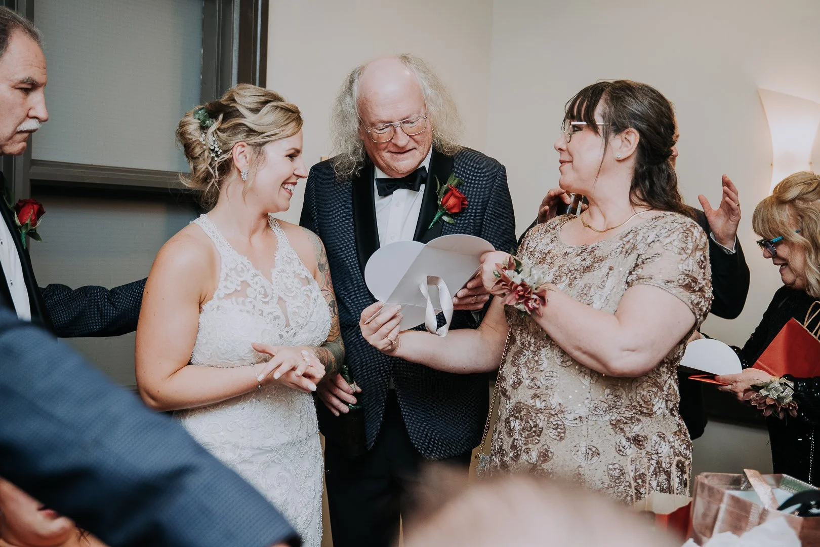 A bride and groom at their wedding surrounded by family members, with one woman reading a note, all smiling.