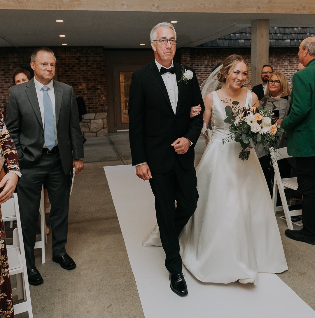 A bride with a veil holding a bouquet of white and peach flowers walking down the aisle accompanied by an older man in a tuxedo, with guests standing on either side.