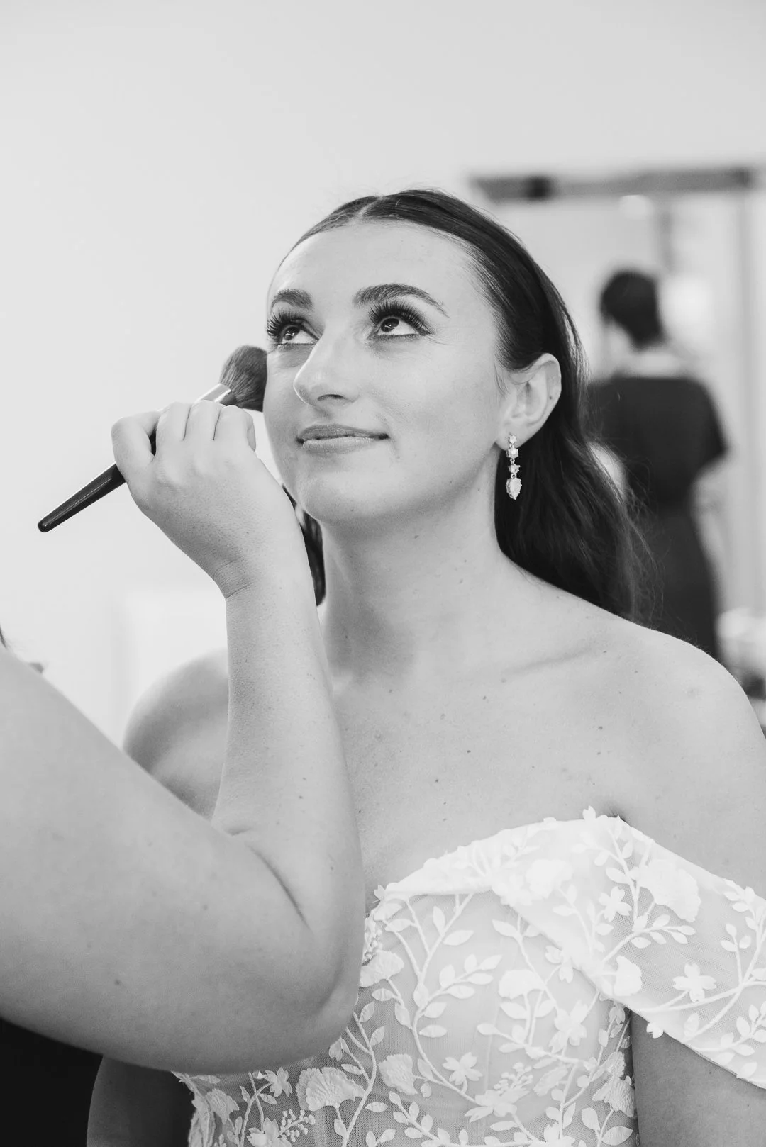 A woman getting her makeup done, looking up and smiling slightly, wearing floral off-shoulder attire and earrings.