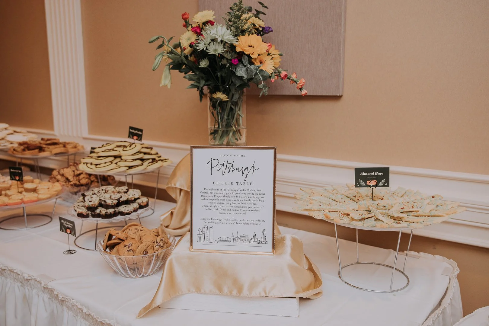 A cookie table with assorted cookies on display, a framed sign about Pittsburgh's cookie history, and a bouquet of flowers in the background.