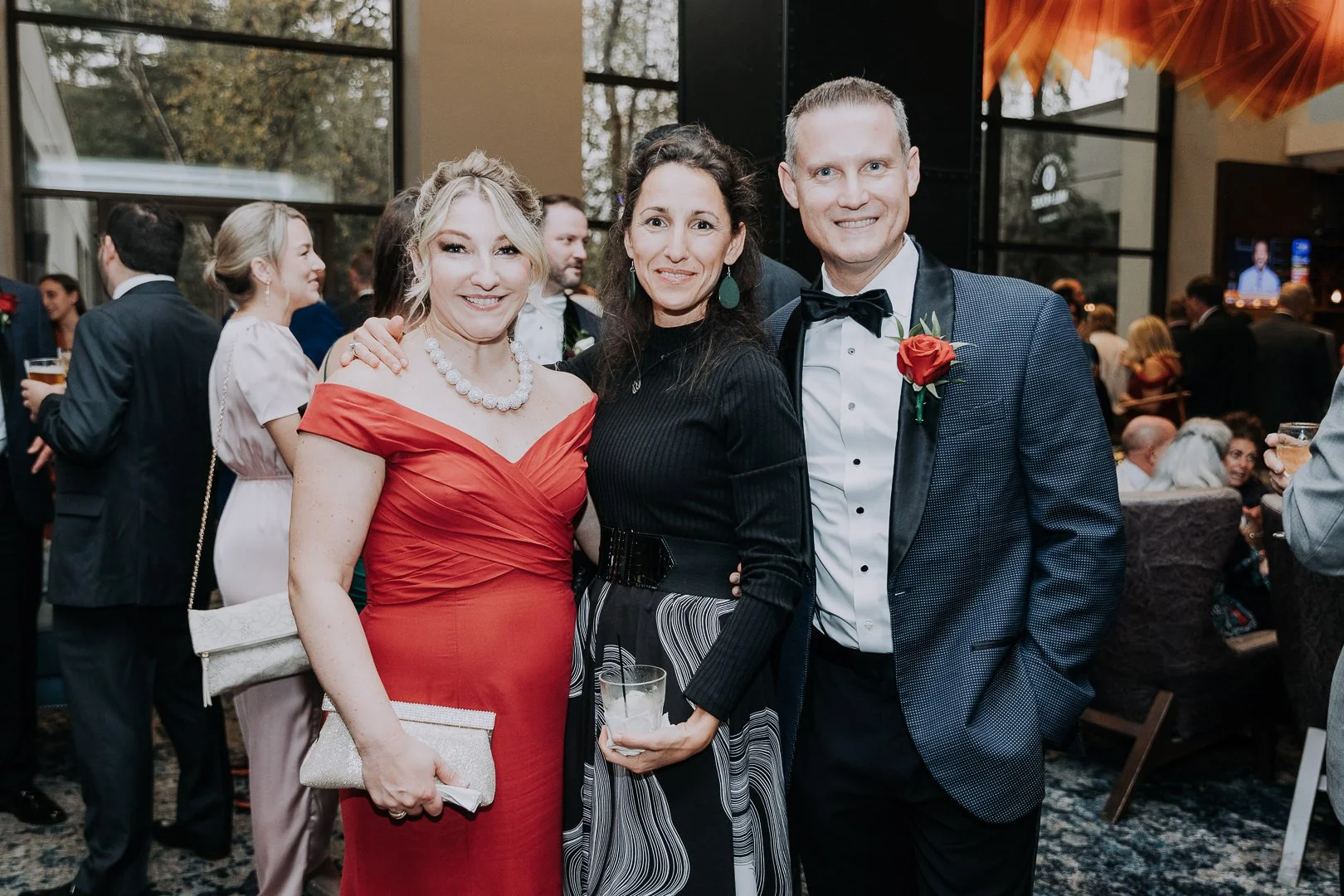 Three people dressed in formal attire at a social event, with a woman in a red dress and three people smiling for the camera in a crowded, elegant setting.