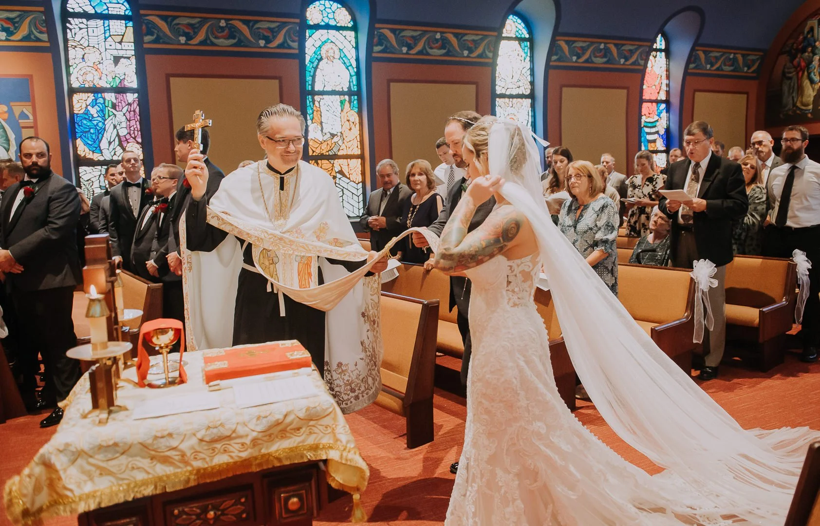 A wedding ceremony inside a church with a bride, groom, and priest. The bride is wearing a lace wedding dress and veil, and the groom is in a suit. The priest is holding a cross and wearing robes. Guests are seated and standing behind them, watching 