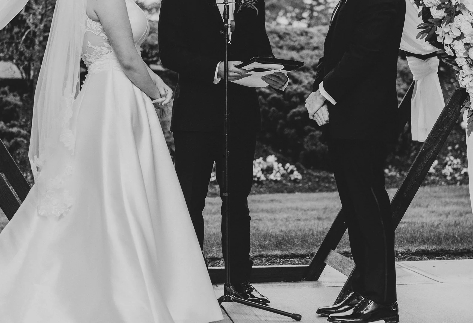 A black and white photo of a wedding ceremony showing a bride in a wedding gown, a groom in a suit, and an officiant holding a tablet or notebook, under an outdoor arch decorated with flowers.