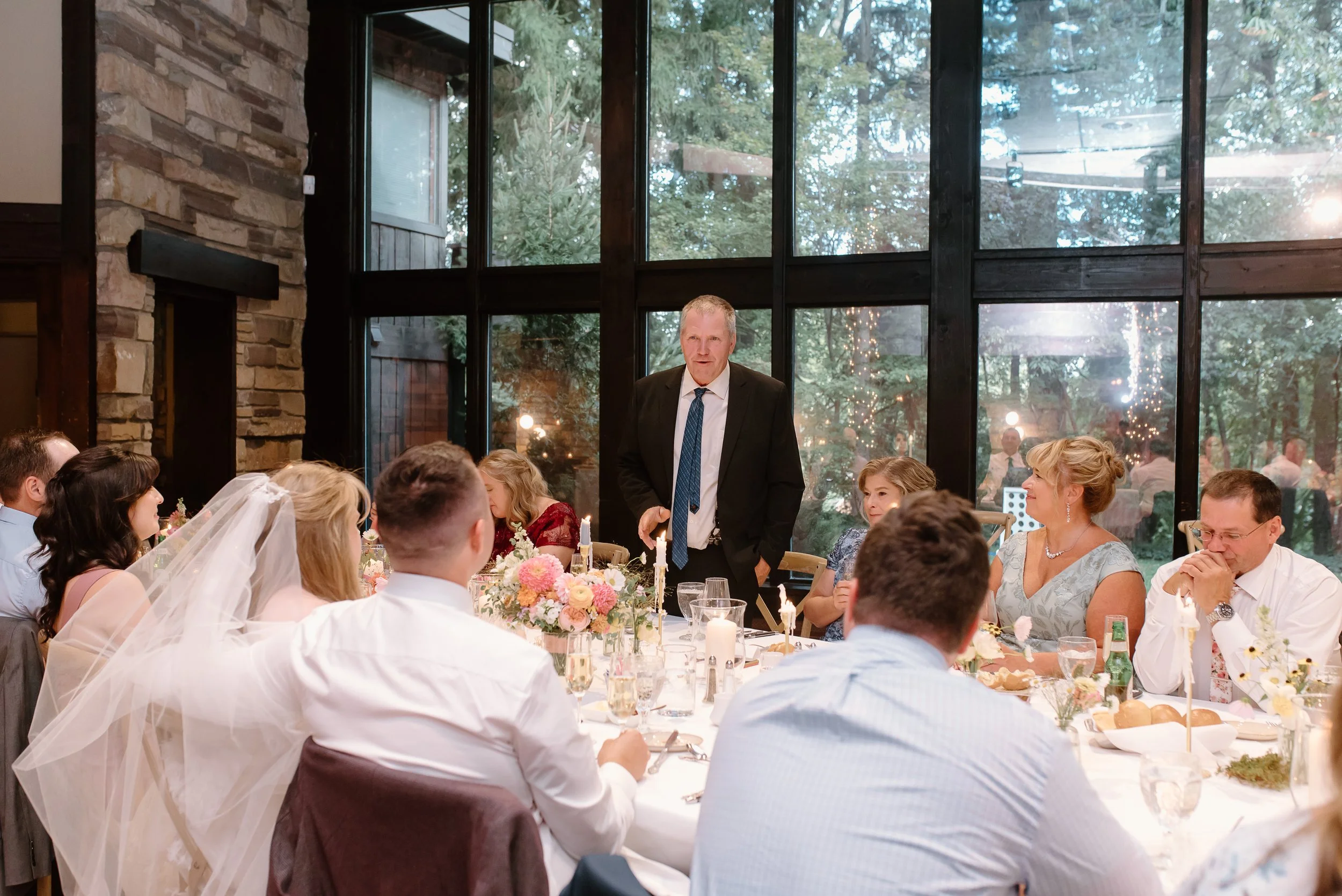 A wedding reception with guests seated around a table, listening to a man in a suit giving a speech, inside a venue with large glass windows and outdoor trees visible.