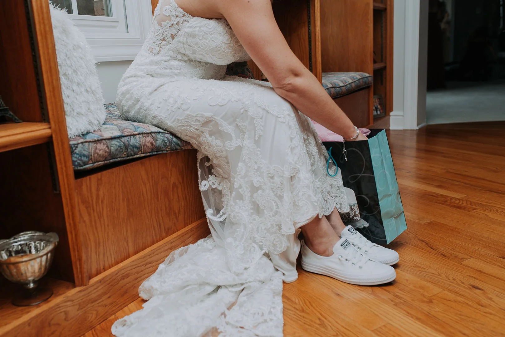 A bride in a lace wedding dress seated on a wooden bench, putting on white sneakers, with a shopping bag beside her on a hardwood floor.