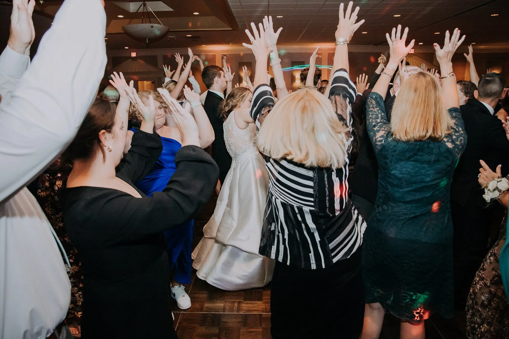 People dancing and celebrating at a wedding reception, with arms raised in the air and a bride in a white gown in the background.