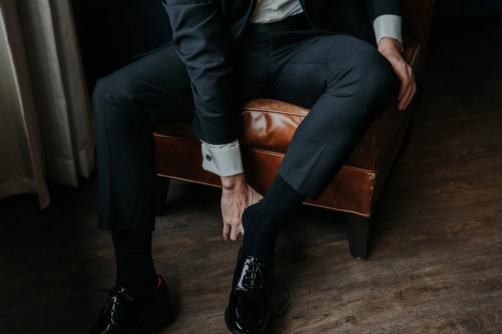 A person dressed in formal attire, sitting on a leather armchair, adjusting their polished black dress shoes.