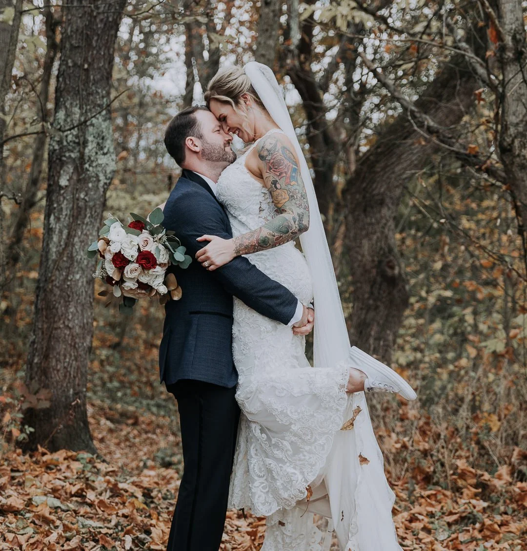 A newlywed couple in a wooded outdoor setting, with the groom lifting the bride and both smiling, the bride holding a bouquet of white and red roses, fall foliage on the ground.