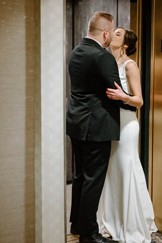 A couple dressed in formal attire sharing a kiss in an elevator, with the woman wearing a white gown and the man in a black suit.