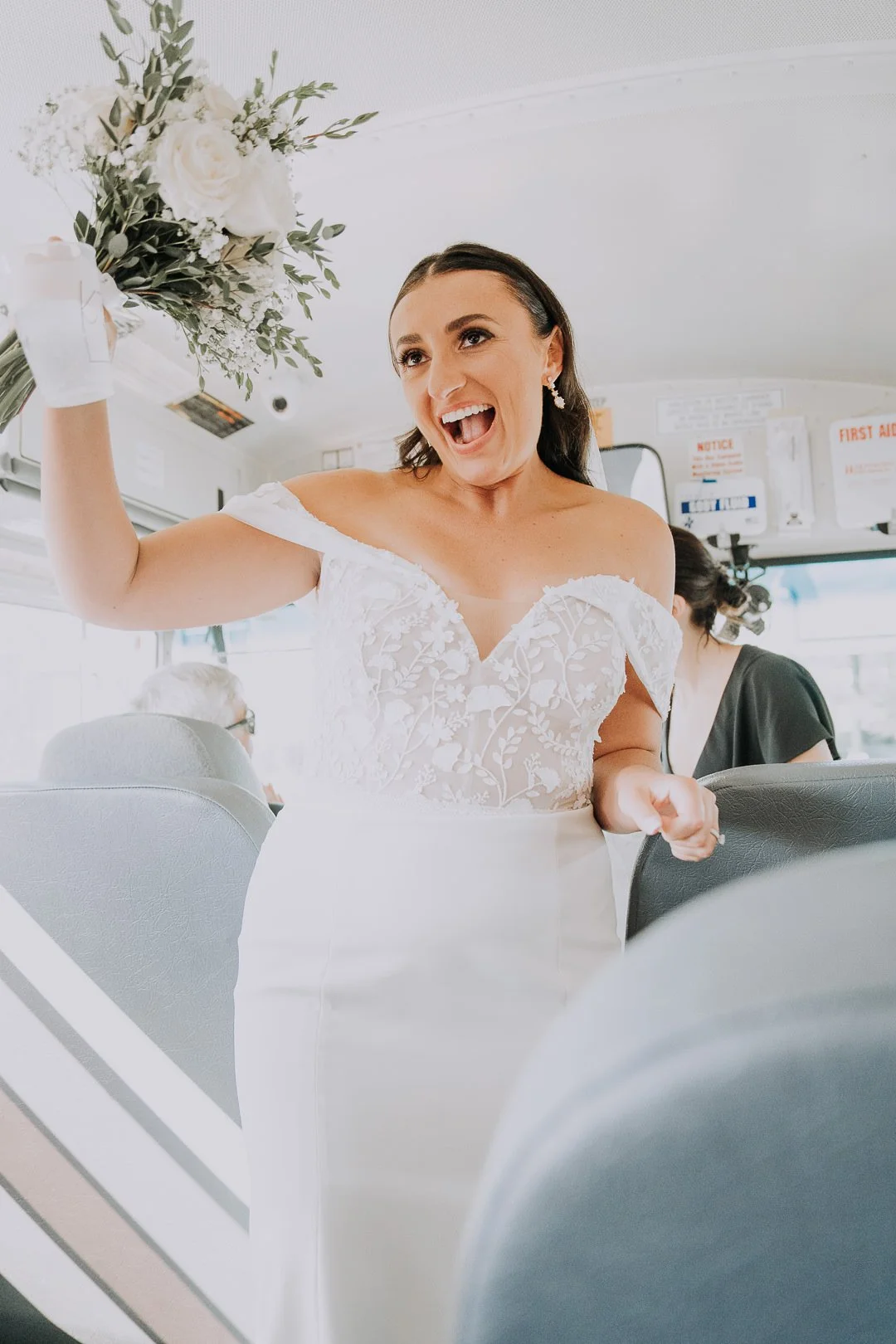 A woman in a wedding dress holding a bouquet of white flowers aboard a bus, smiling and appearing joyful.