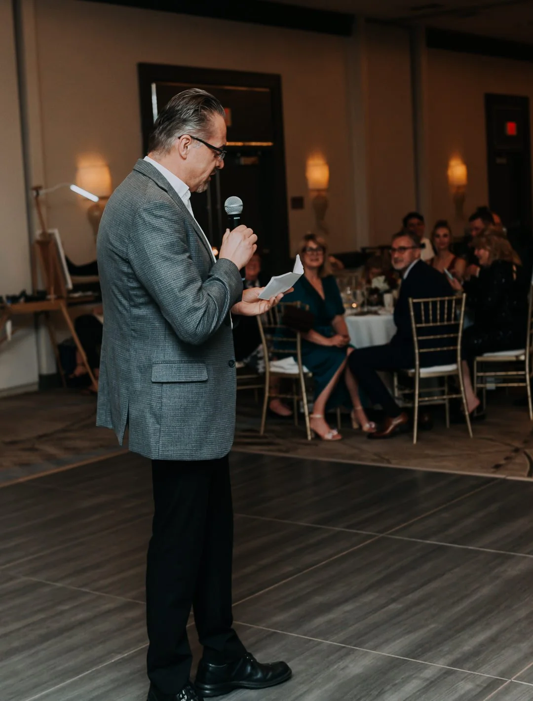 A man in a gray blazer and black pants standing on a wooden floor, reading from a small piece of paper into a microphone at a formal event with guests seated at round tables in the background.