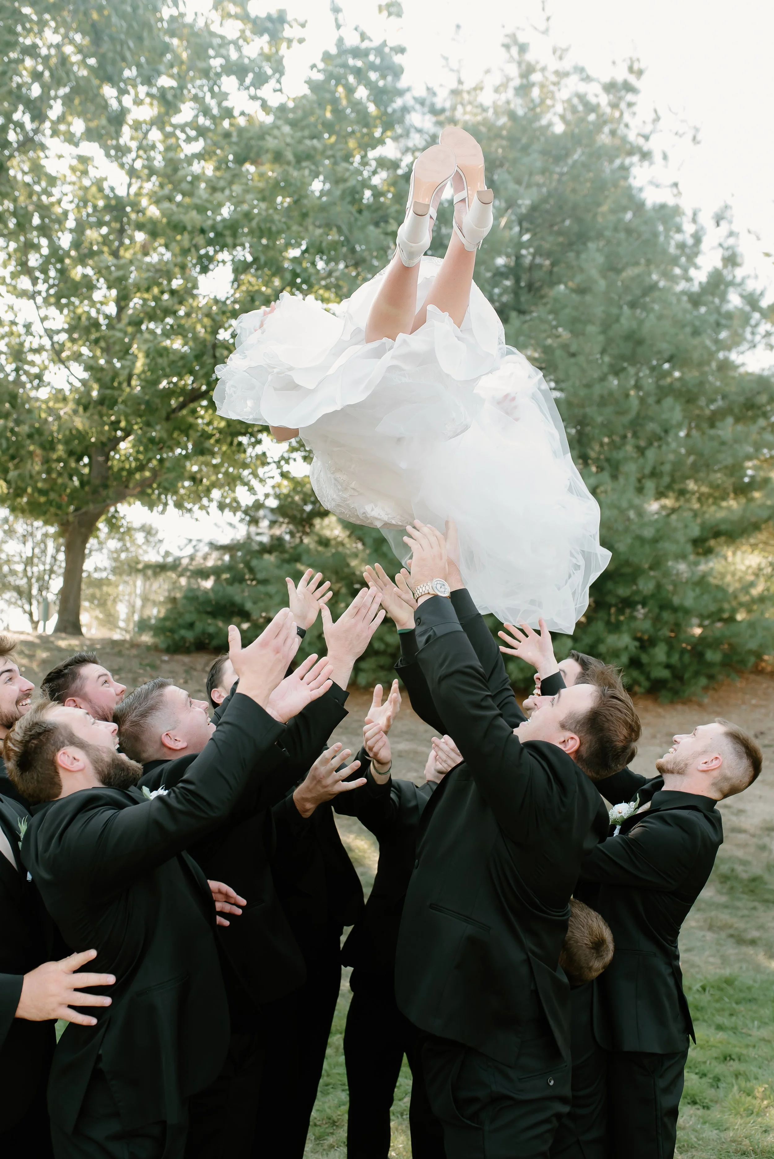 A bride in a white wedding dress being tossed into the air by a group of men in black suits outdoors on a sunny day.