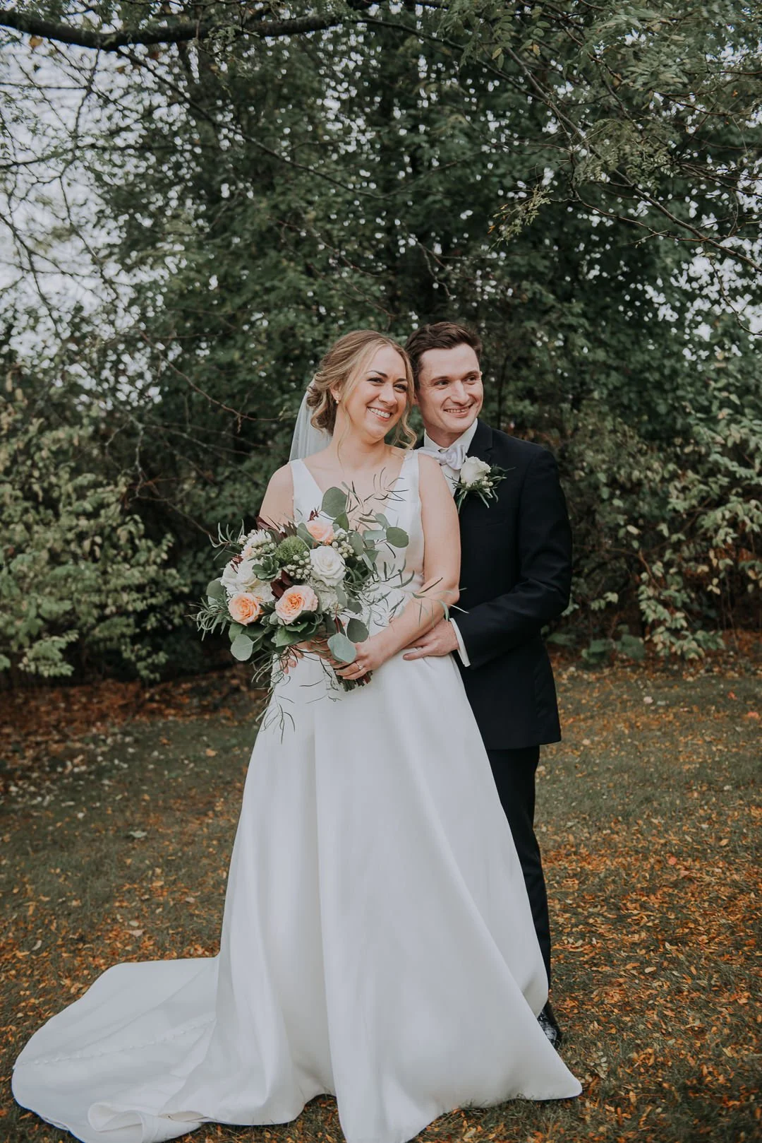 A bride and groom standing outdoors on a wedding day, smiling and looking happy. The bride is holding a bouquet of flowers, and they are surrounded by trees and greenery.