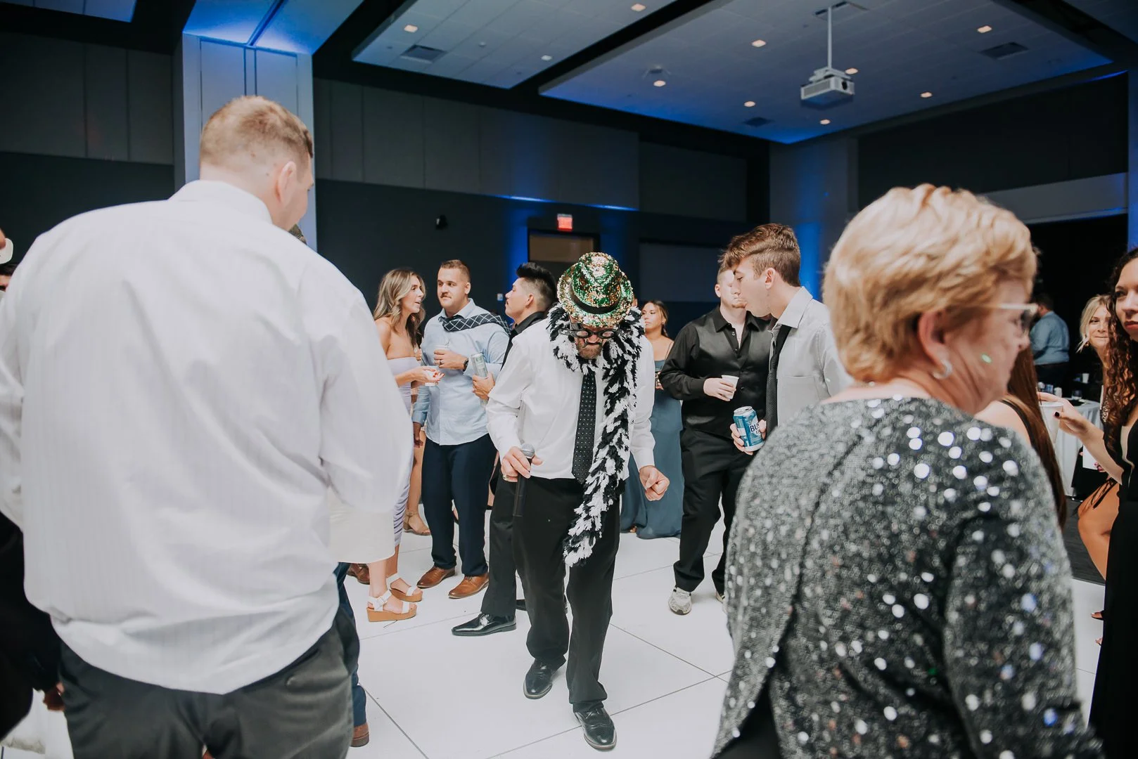 People dancing and socializing at a festive event indoors, with a man wearing a party hat, scarf, glasses, and suit in the center.