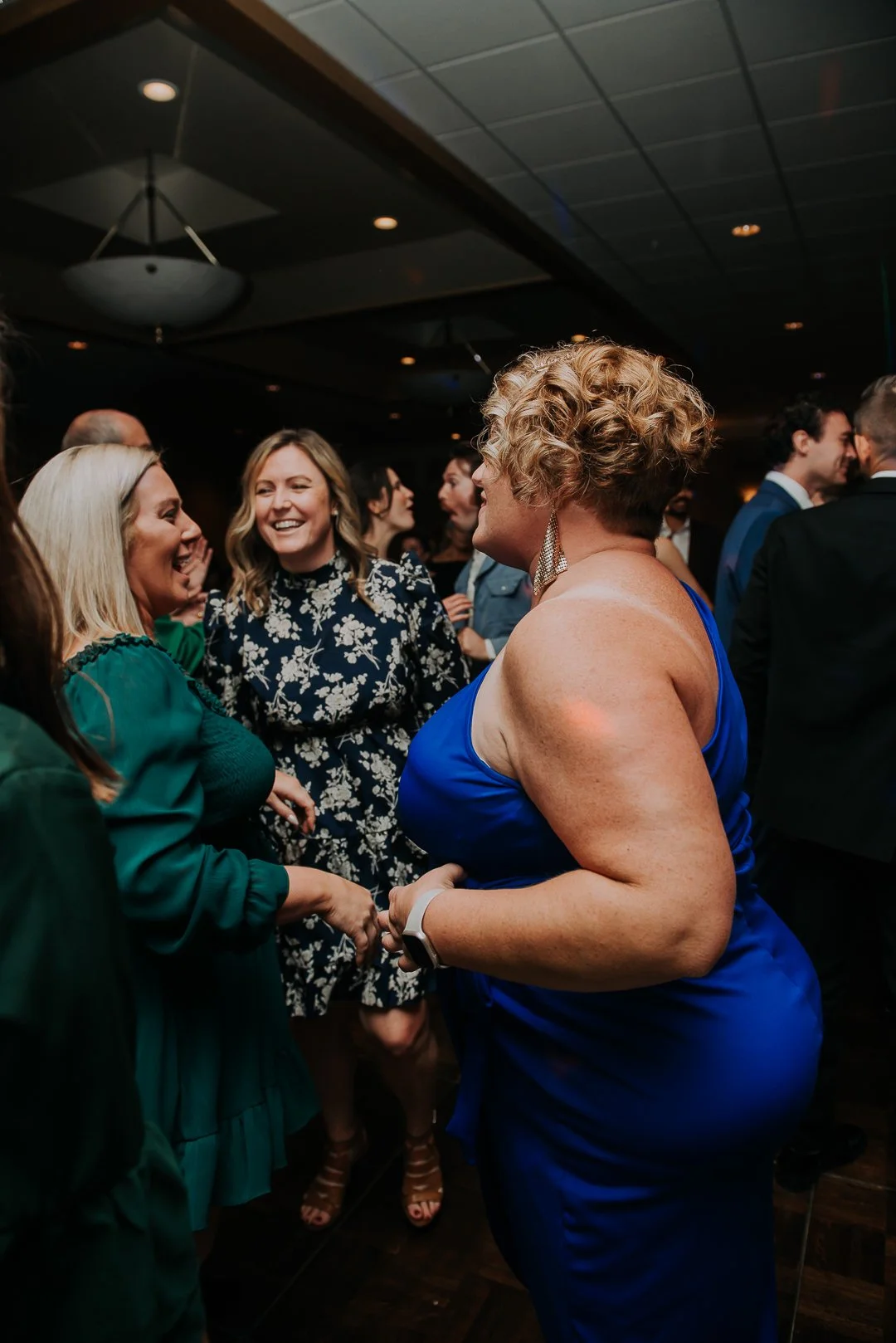 Group of women smiling and chatting at an indoor social event, with dim lighting, dark ceiling, and other guests in the background.