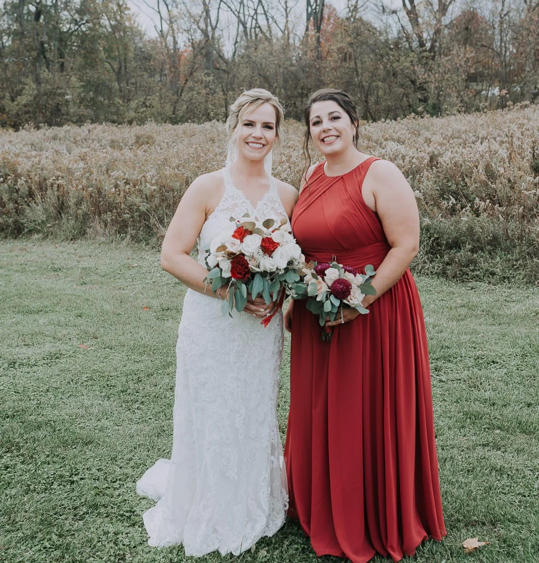 Two women standing outside in a grassy field, smiling and holding bouquets of flowers. One woman is in a white lace wedding dress with blonde hair, and the other in a sleeveless red gown with dark hair. Trees with fall foliage are in the background.