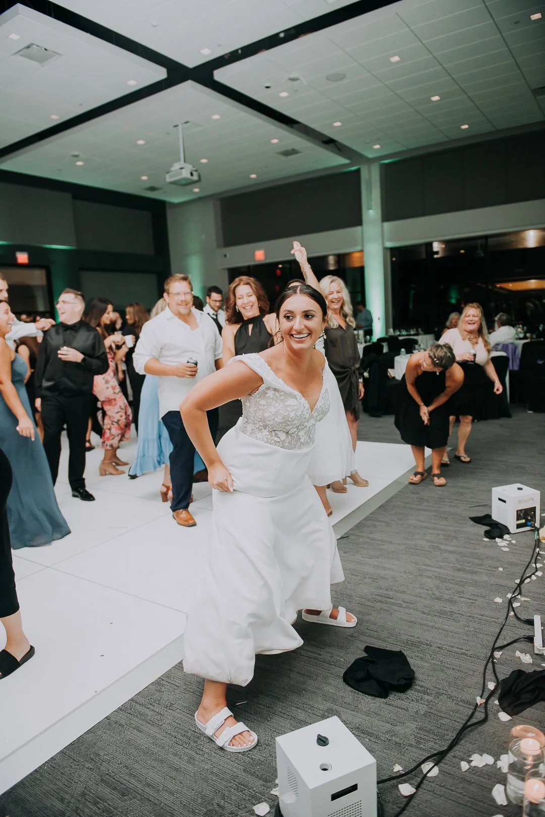 A woman in a white wedding dress dancing and smiling at a wedding reception with guests in the background.