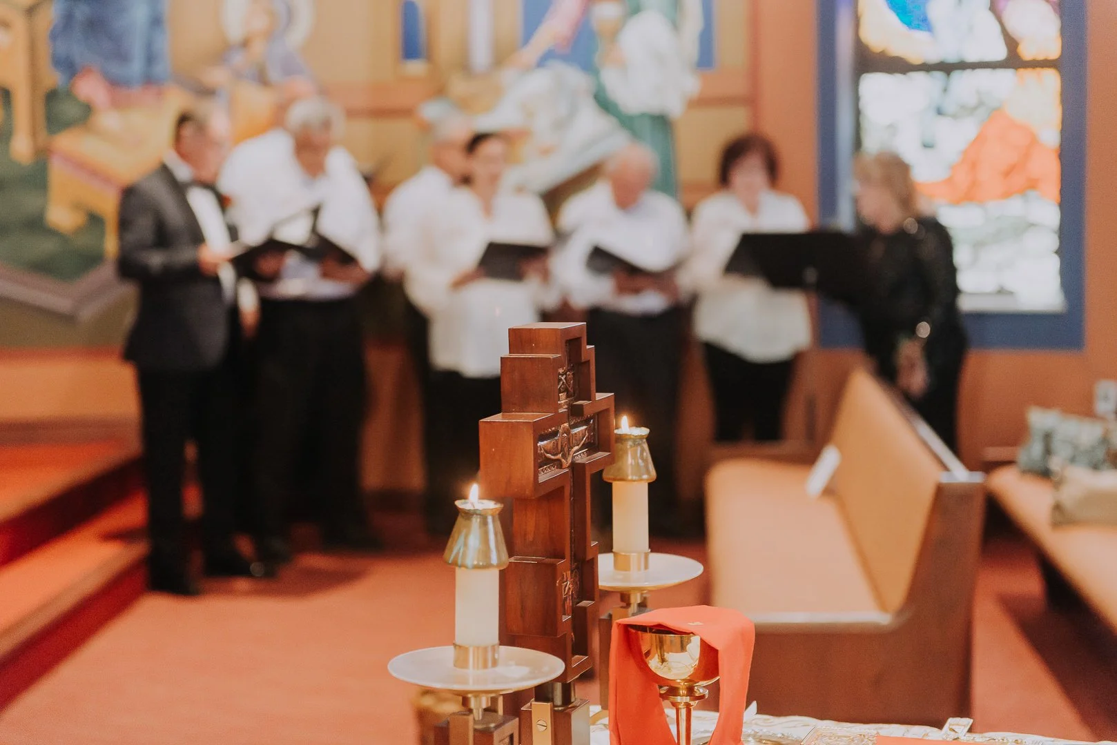 A church setting with a group of people, some holding hymn books, in the background. In the foreground, there is a wooden cross and two lit candles on stands, as well as an orange cloth draped over one candle holder.