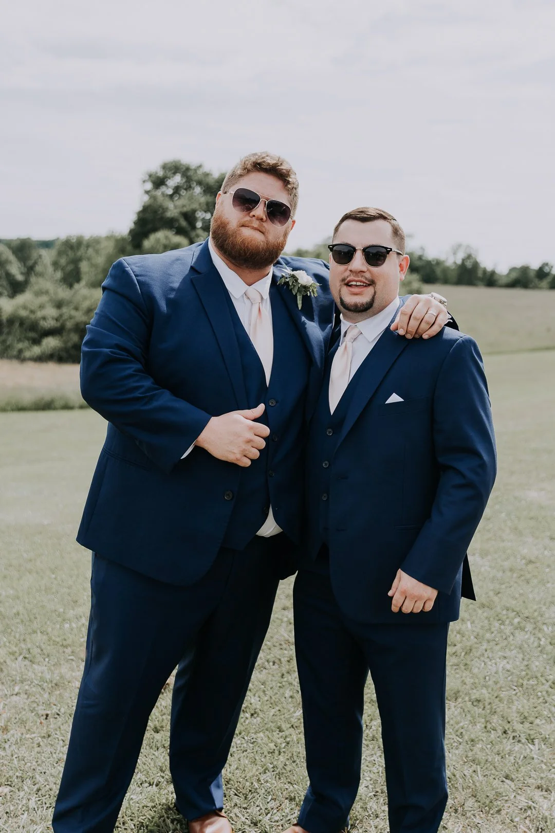 Two men in blue suits and sunglasses standing outdoors with arms around each other, smiling at the camera, on a grassy field with trees in the background.