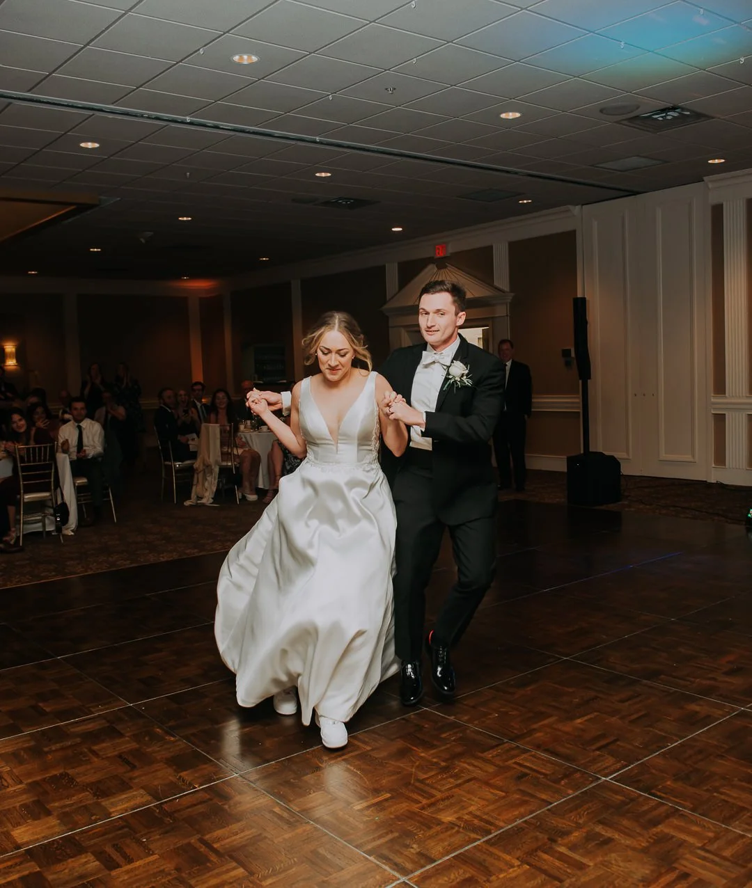 Couple in wedding attire dancing at a wedding reception with seated guests in the background.