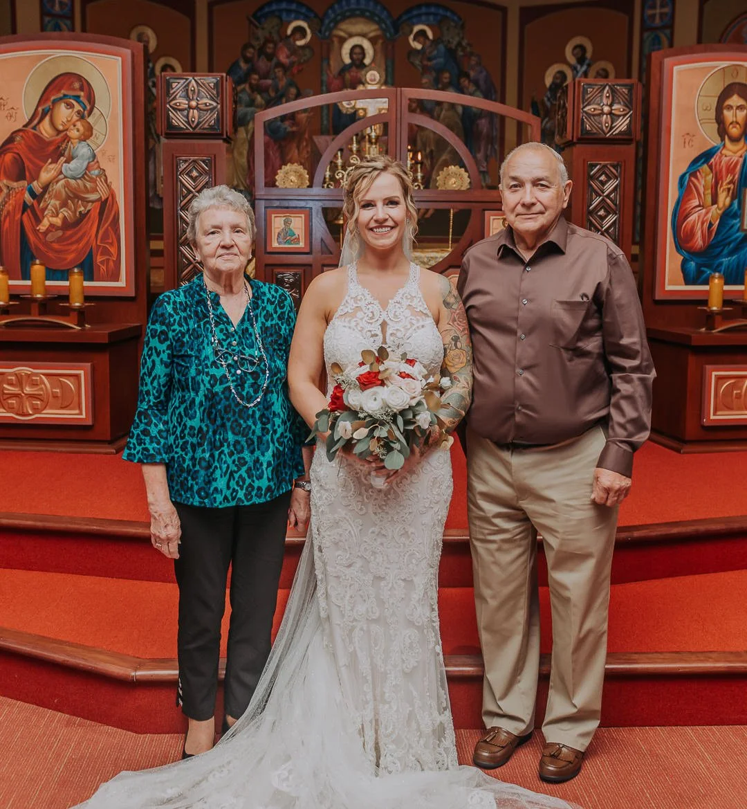A bride in a white lace wedding dress holding a bouquet stands with an elderly woman in a blue leopard print blouse and a necklace, and an older man in a brown dress shirt and khakis inside a church with religious icons and artwork in the background.