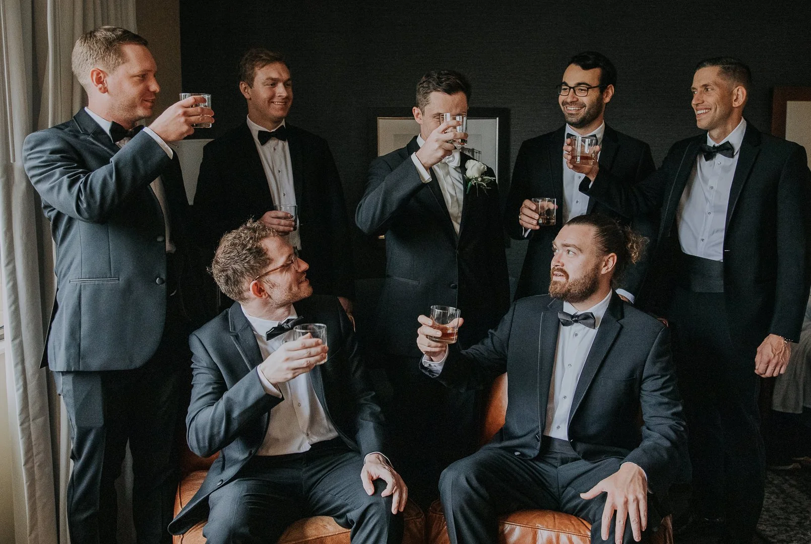 Group of seven men in suits and tuxedos having drinks and toasting at a celebration or wedding reception in a dimly lit room.