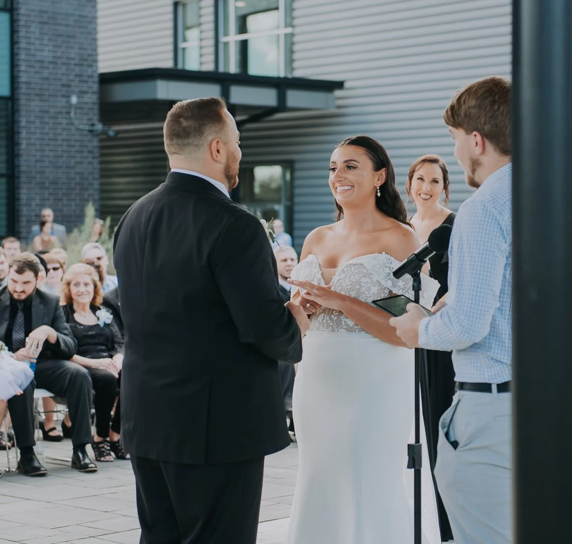 A couple getting married at an outdoor wedding ceremony, with guests seated and smiling in the background.