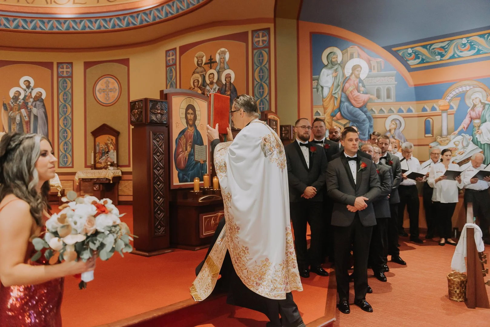 A wedding ceremony inside a church with a priest reading from a red book, with the bride holding a bouquet of flowers in the foreground, and the groom and groomsmen standing in a line, all dressed in formal attire, in front of religious iconography a